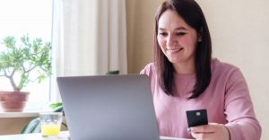 A smiling woman sitting in front of her laptop with a credit card holding in her hand to make a payment